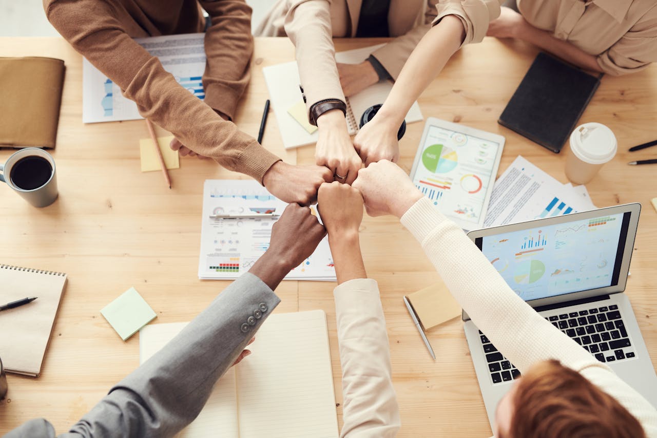 why-choose-us Top view of a diverse team fist bumping over a meeting table with paperwork and laptops, symbolizing teamwork.